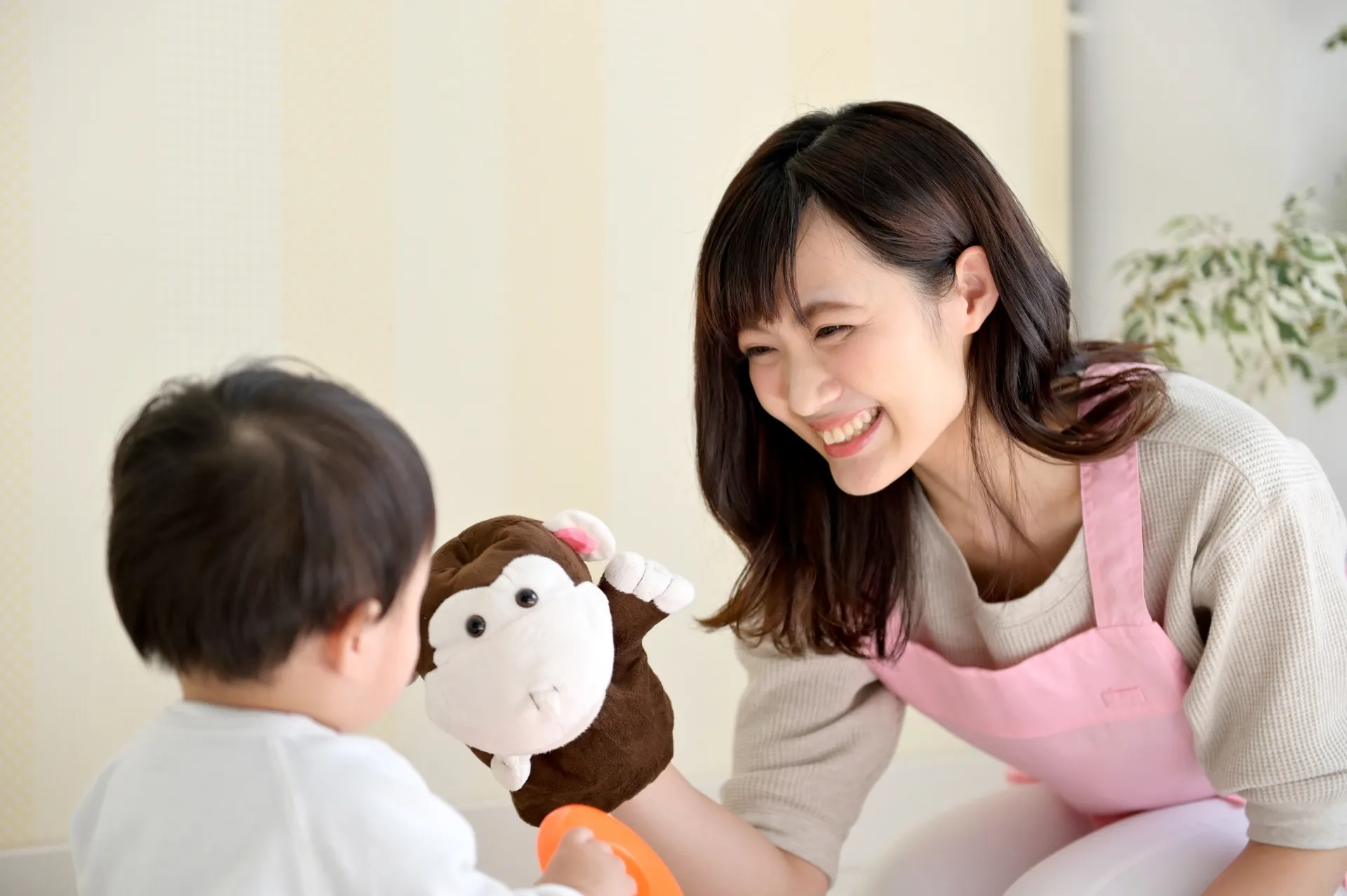 ピンクのエプロンを着たシッターが人形で子どもと遊ぶ様子